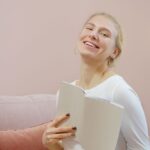 Vrolijke vrouw in het wit die op een pastelbank een boek vasthoudt en warmte en vreugde uitstraalt